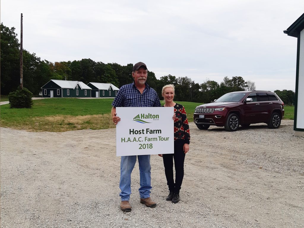 Deerfield Farm owners holding a sign about hosting a farm tour in 2018, standing at their farm entrance